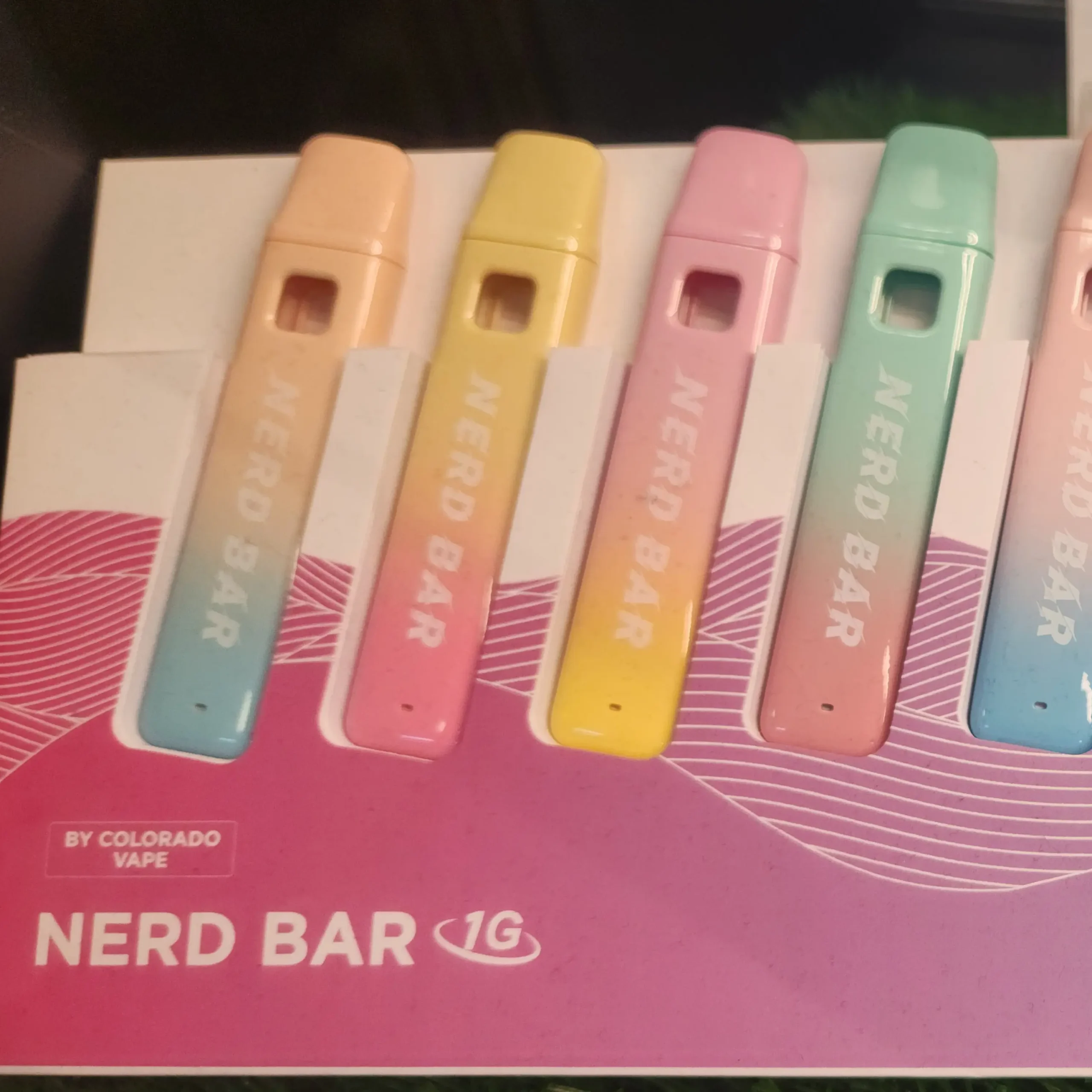 A display of colorful Nerd Bar vape pens in pastel shades, labeled with "NERD BAR" on a pink background.